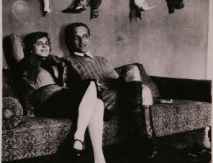Fotografia czarnobiała. Na kanapie siedzi uśmiechnięta młoda kobieta obok młodego mężczyzny. Nad nimi, na ścianie powieszone trofea myśliwskie.]