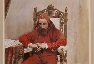 Kolorowa fotografia przedstawiająca mężczyznę w stroju Stańczyka siedzącego na ozdobionym drewnianym krześle. Czerwony strój zawiera w sobie również czerwone nakrycie głowy.
