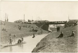 Fotografia czarnobiała. Osoby odpoczywające na rzeką. Część osób znajduje się na brzegu, część w łódce na wodzie. W tle most.