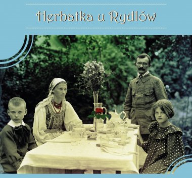 kolorowa grafika. Na niebieskim tle napis: Herbatka u Rydlów. Pośrodku czarno-białą fotografia. Rodzina siedząca przy stole, w tle zieleń. Na stole dzbanek herbaciany, filiżanki i kwiaty.