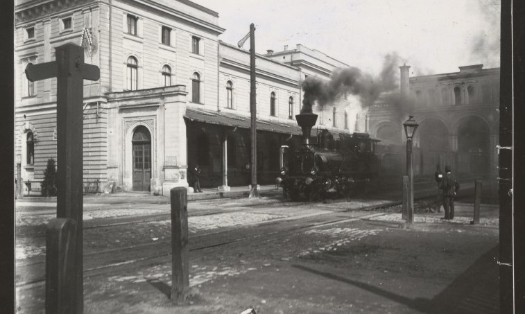 Czarno biała fotografia lokomotywy wjeżdżającej na stację.