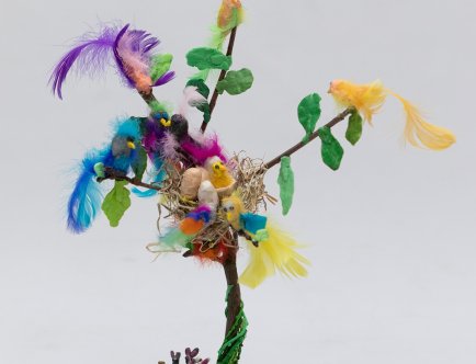 Fotografia drzewka emausowego. Na gałęziach kolorowe piórka. W środku drzewka gniazdo z ptaszkami.