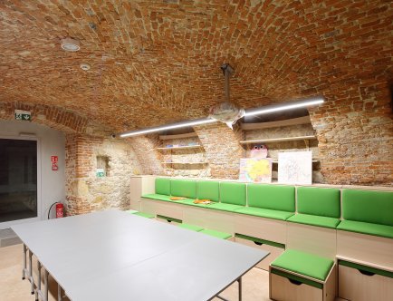Kolorowa fotografia przedstawiająca wnętrze lokalu. Duży stół oraz siedziska ustawione wzdłuż ściany.. W ścianie półki.