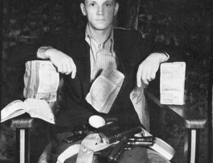 Propagandowa fotografia czarnobiała. Mężczyzna siedzi na krześle. Na kolanach i pod rękami ma pliki banknotów, stułę, brewiarz oraz pistolet.