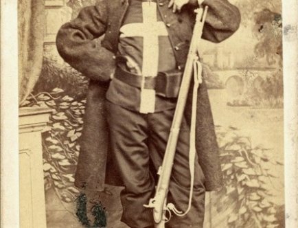 Zdjęcie w sepii przedstawiające mężczyznę w oficjalnym stroju trzymającym strzelbę.