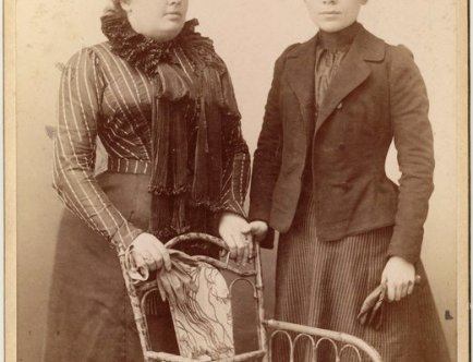 Zdjęcie w sepii. Dwie kobiety w sukniach i kapeluszach stojące za krzesłem.