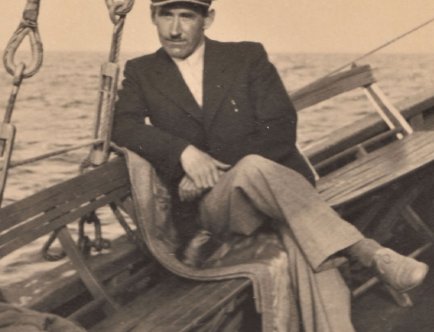 Na zdjęciu przedstawiony jest elegancko ubrany mężczyzna w berecie na głowie siedzący na ławce na pokładzie statku.
