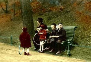 Fotografia kolorowa. Starszy Żyd z dwójką chłopców siedzi na ławce. Obok bawią się dwie dziewczynki. W tle skarpa z drzewami