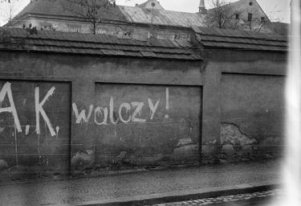 Fotografia czarnobiała. Od lewej strony, na murze, duży, ręcznie napisany białą farbą napis  „AK walczy”