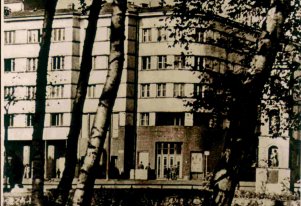 Fotografia czarnobiała. Budynek od strony Parku Krakowskiego w ujęciu między drzewami.