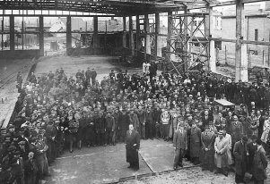 Fotografia czarnobiała zrobiona z podwyższenia w hali fabrycznej bez okien. Na pierwszym planie Oskar Schindler. Wkoło niego duża grupa pracowników