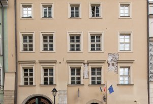 Kolorowa fotografia przedstawiająca wnętrze jednego z pokoi domu mieszczańskiego. Na środku stół wraz z krzesłami, przy ścianach drewniane meble, na ścianach obrazy.