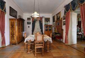 Kolorowa fotografia przedstawiająca wnętrze jednego z pokoi domu mieszczańskiego. Na środku stół wraz z krzesłami, przy ścianach drewniane meble, na ścianach obrazy.